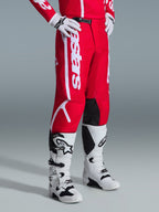 Alpinestars 2026 Fluid Apex Pants, spodnie motocrossowe, czerwono-białe, noszone z biało-czarnymi butami i rękawicami motocrossowymi, z wyraźnym białym brandingiem i czarnymi panelami na wewnętrznej stronie kolan, zaprojektowane dla wydajności off-road