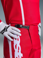 Alpinestars 2026 Fluid Apex Pants, spodnie motocrossowe, czerwono-białe, zbliżenie na czarny system zapięcia zapadkowego w pasie, noszone z białymi rękawicami Alpinestars i dopasowaną czerwoną koszulką, wytrzymała odzież wyczynowa do jazdy off-road.