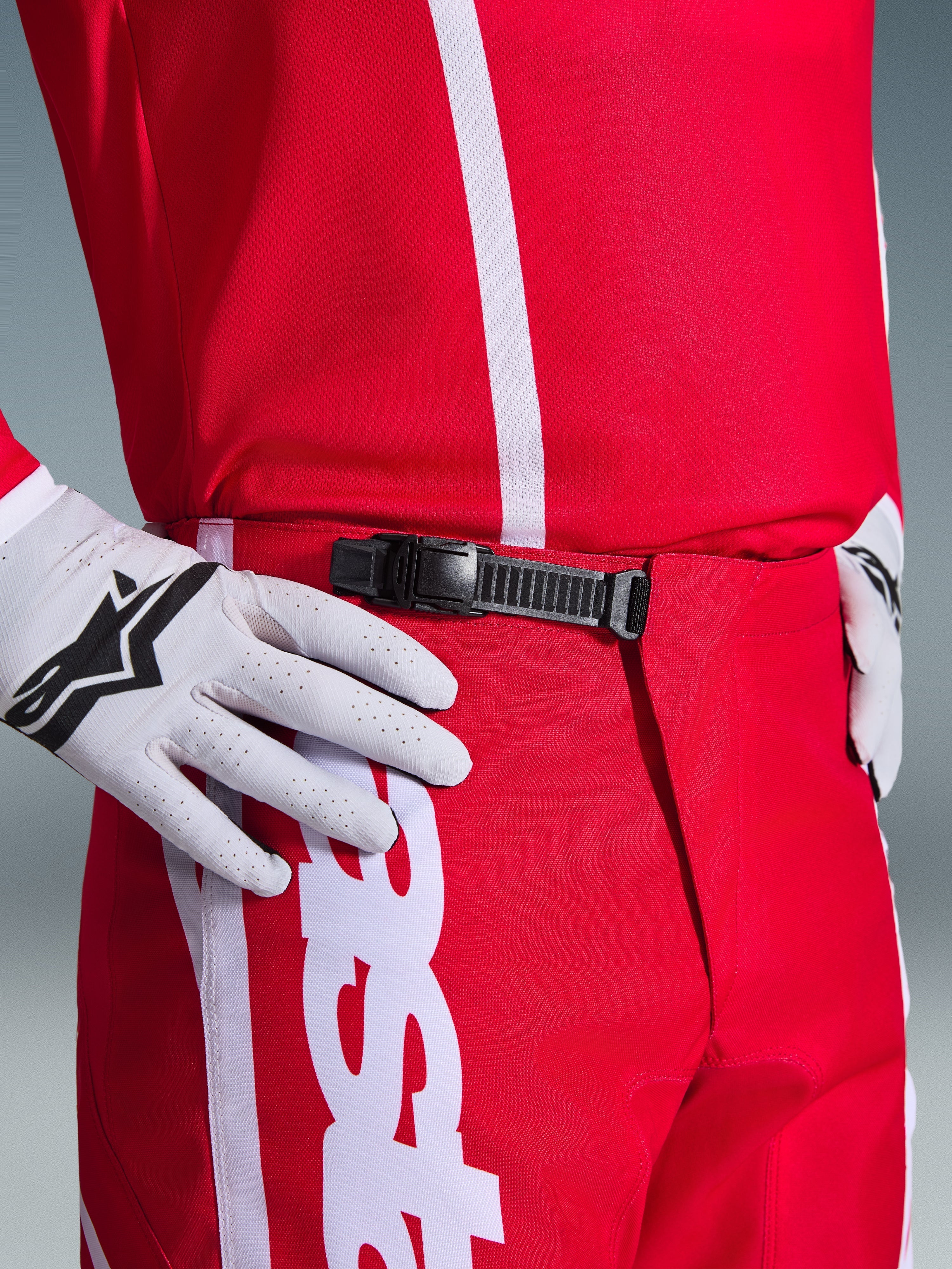 Alpinestars 2026 Fluid Apex Pants, spodnie motocrossowe, czerwono-białe, zbliżenie na czarny system zapięcia zapadkowego w pasie, noszone z białymi rękawicami Alpinestars i dopasowaną czerwoną koszulką, wytrzymała odzież wyczynowa do jazdy off-road.