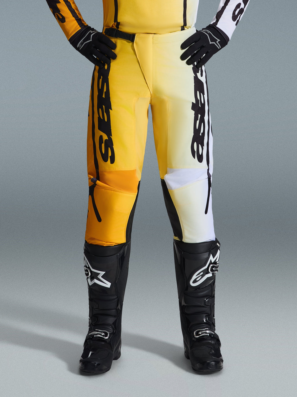 Alpinestars 2026 Fluid Apex Pants, spodnie motocrossowe, żółto-czarne, dwukolorowy design z żółtymi i białymi panelami, wyraźne pionowe logo „astars”, noszone z czarnymi butami i rękawicami motocrossowymi, odzież wyczynowa do jazdy terenowej