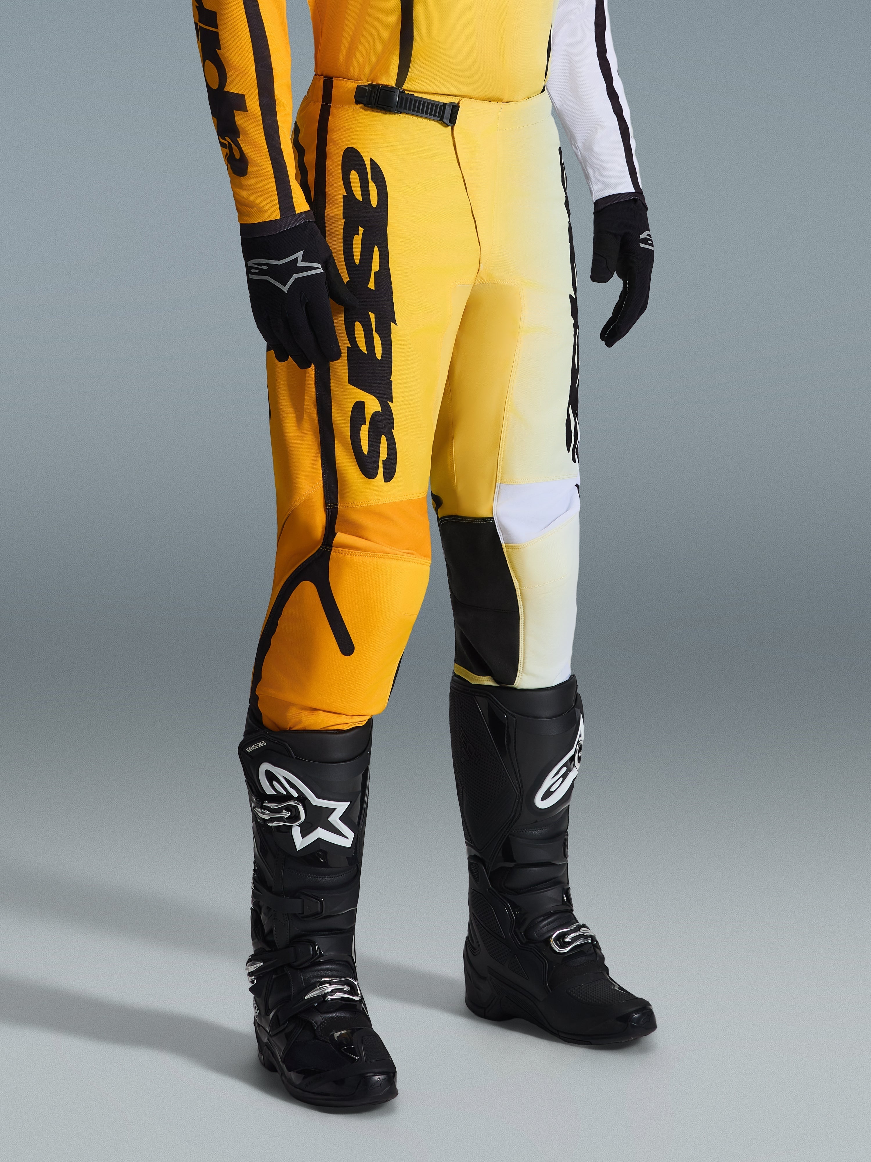 Alpinestars 2026 Fluid Apex Pants, spodnie motocrossowe, żółto-czarne, noszone z czarnymi butami i rękawicami, z wyrazistym brandingiem „astars” na udzie, ergonomiczna konstrukcja ze wzmocnionymi panelami, wytrzymały materiał do jazdy terenowej