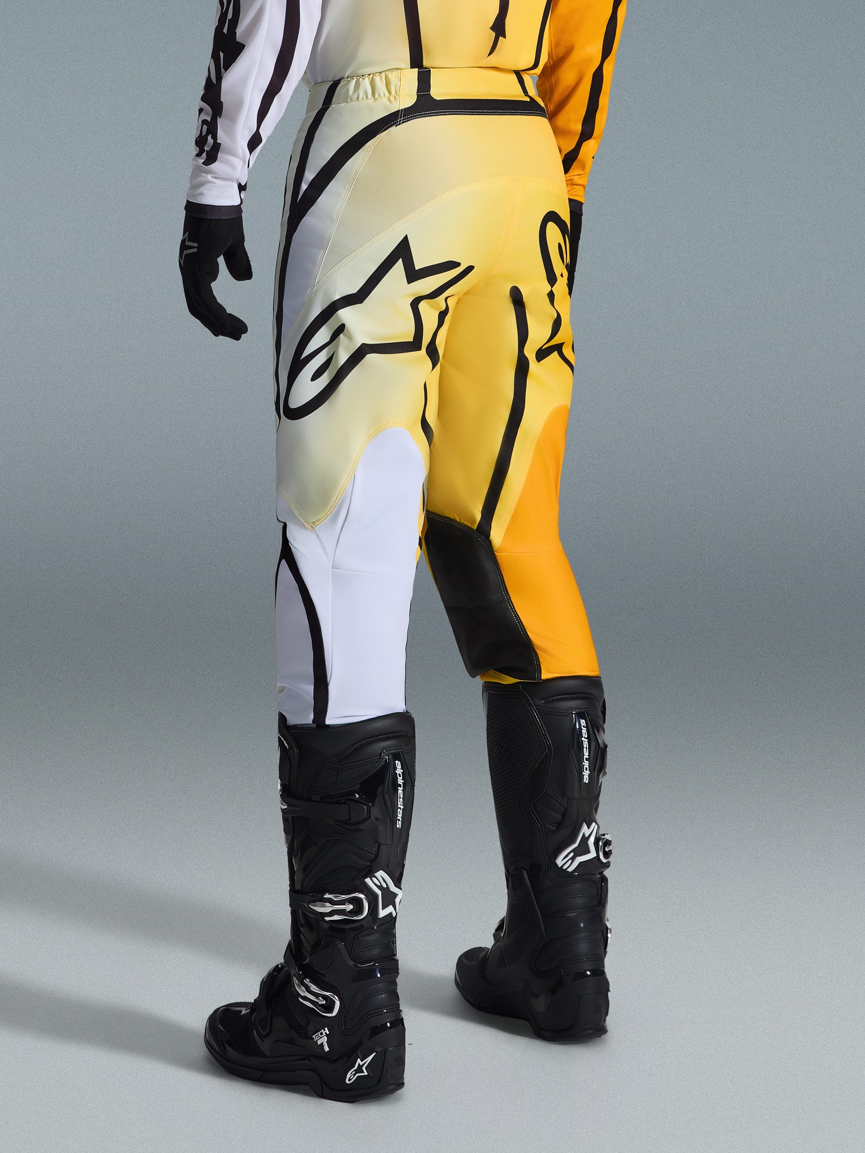 Alpinestars 2026 Fluid Apex Pants, spodnie motocrossowe, żółto-czarny gradient z białymi i pomarańczowymi akcentami, widok z tyłu, noszone z pasującą koszulką i czarnymi butami Tech 7, wyraźne czarne logo Alpinestars na siedzeniu, profesjonalna odzież do jazdy off-road