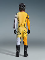 Alpinestars 2026 Fluid Apex Pants, spodnie motocrossowe, żółto-czarne, widok od tyłu motocyklisty w pełnym stroju motocrossowym, pasująca koszulka z białymi i pomarańczowymi gradientami, czarny kask, czarne buty i czarne rękawice, skoordynowany zestaw off-road