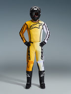 Alpinestars 2026 Fluid Apex Pants, spodnie motocrossowe w kolorze żółtym, czarnym i białym, noszone jako kompletny zestaw wyścigowy z dopasowaną koszulką z gradientem, czarnym kaskiem i butami, profesjonalna odzież do jazdy off-road.