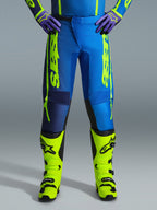 Alpinestars 2026 Fluid Apex Pants, spodnie motocrossowe, niebieskie i żółte fluo, noszone z pasującymi żółtymi butami fluo i fioletowymi rękawicami, wyraźne żółte logo na bokach, ergonomiczna konstrukcja do wyścigów terenowych