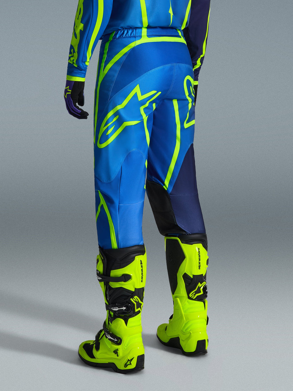 Alpinestars 2026 Fluid Apex Pants, spodnie motocrossowe, niebieskie i żółte fluo, widok od tyłu na motocyklistę w pasującym stroju i jaskrawożółtych butach, wyraziste logo Alpinestars, ergonomiczny design