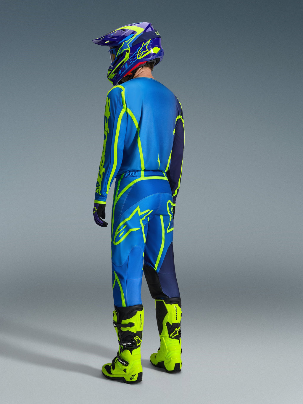 Zawodnik motocrossowy widziany od tyłu w spodniach Alpinestars Fluid Apex 2026 w kolorze niebieskim i żółtym fluo, dopasowanej niebiesko-żółtej koszulce i kasku oraz jaskrawych neonowożółtych butach, skoordynowany zestaw wyścigowy do jazdy terenowej