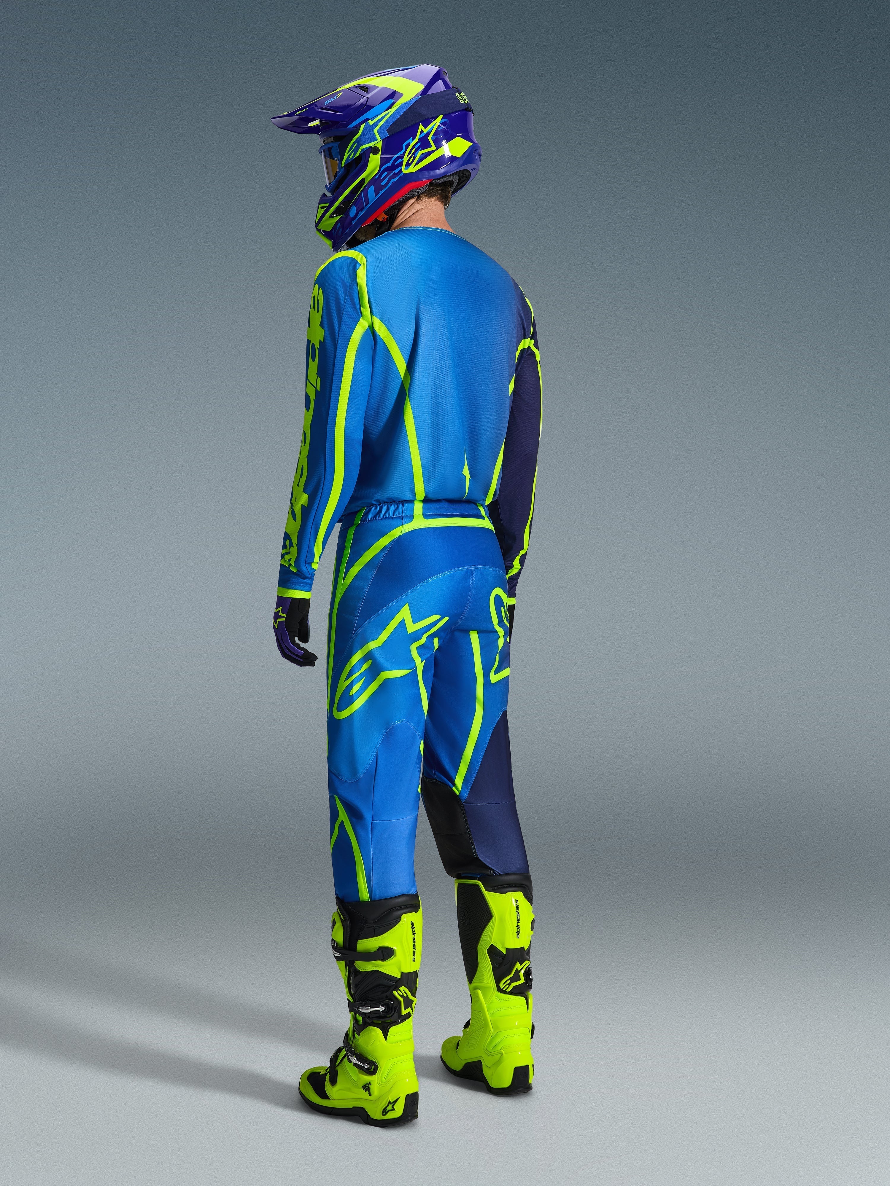 Zawodnik motocrossowy widziany od tyłu w spodniach Alpinestars Fluid Apex 2026 w kolorze niebieskim i żółtym fluo, dopasowanej niebiesko-żółtej koszulce i kasku oraz jaskrawych neonowożółtych butach, skoordynowany zestaw wyścigowy do jazdy terenowej
