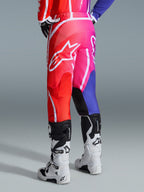 Alpinestars 2026 Fluid Apex Pants, spodnie motocrossowe, wielokolorowe czerwone, różowe i fioletowe, widok z tyłu na motocyklistę w spodniach i biało-czarnych butach motocrossowych, duże białe logo Alpinestars, jaskrawy gradientowy wzór