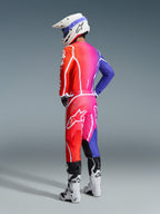 Zawodnik motocrossowy w stroju Alpinestars 2026 Fluid Apex, wielokolorowa bluza i spodnie z gradientem czerwieni, różu i fioletu, białe logo, biały kask i białe buty, dopasowany strój do wyścigów off-road