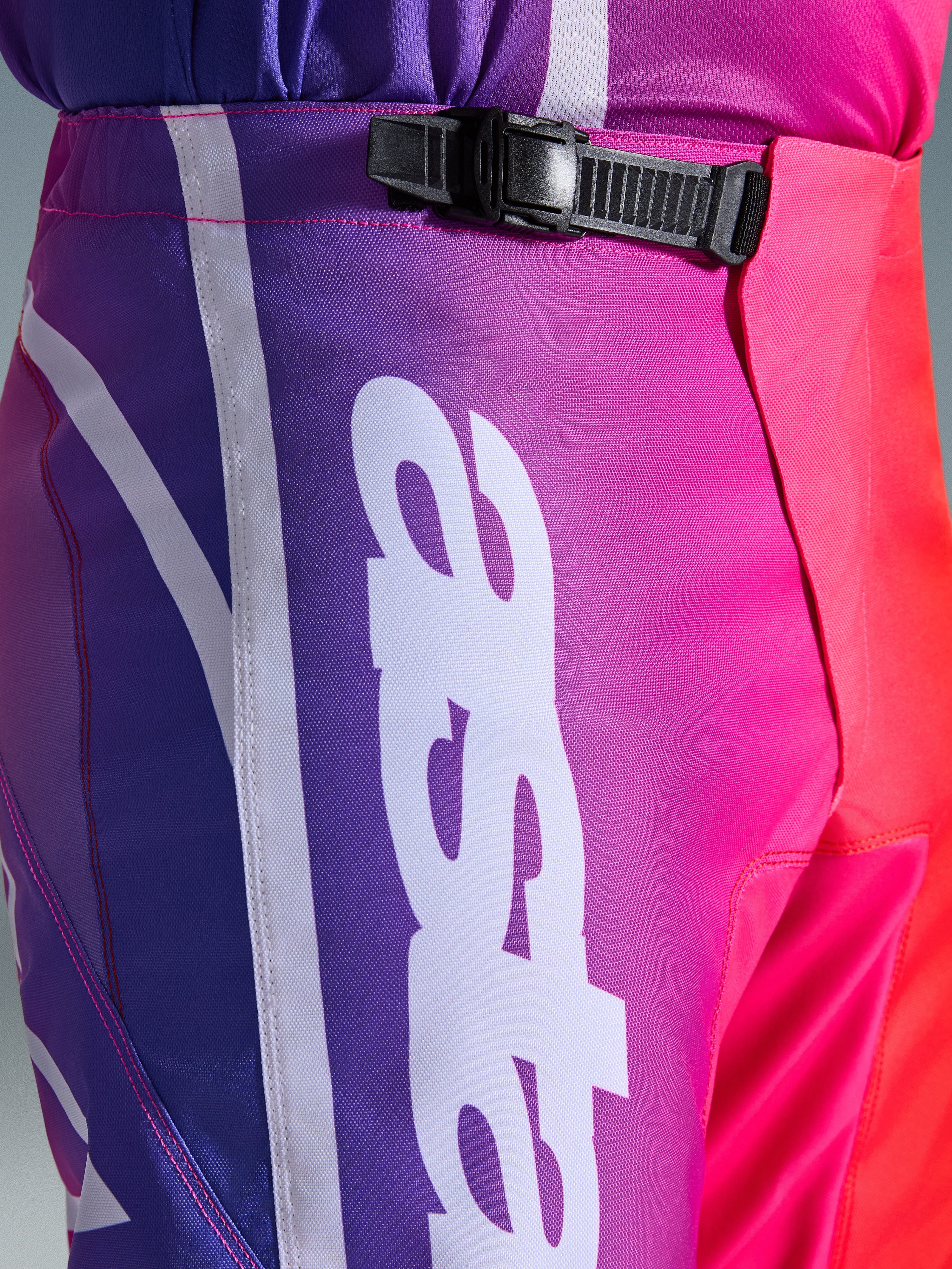 Alpinestars 2026 Fluid Apex Pants, spodnie motocrossowe, wielokolorowe fioletowo-różowo-czerwone, zbliżenie na talię i biodra z czarnym systemem zapięcia zapadkowego i dużym białym pionowym logo Alpinestars, żywy gradientowy wzór z wytrzymałą teksturą materiału
