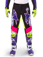 Alpinestars Limited Edition Supertech Dirt Studios Pants, spodnie motocrossowe, białe, fioletowe i żółte fluo, czarne panele z neonowo-żółtymi i różowymi akcentami, duże fioletowe logo gwiazdy, widoczne z pasującymi neonowo-żółtymi rękawicami i fioletowymi butami wyścigowymi, wytrzymała odzież wyścigowa off-road