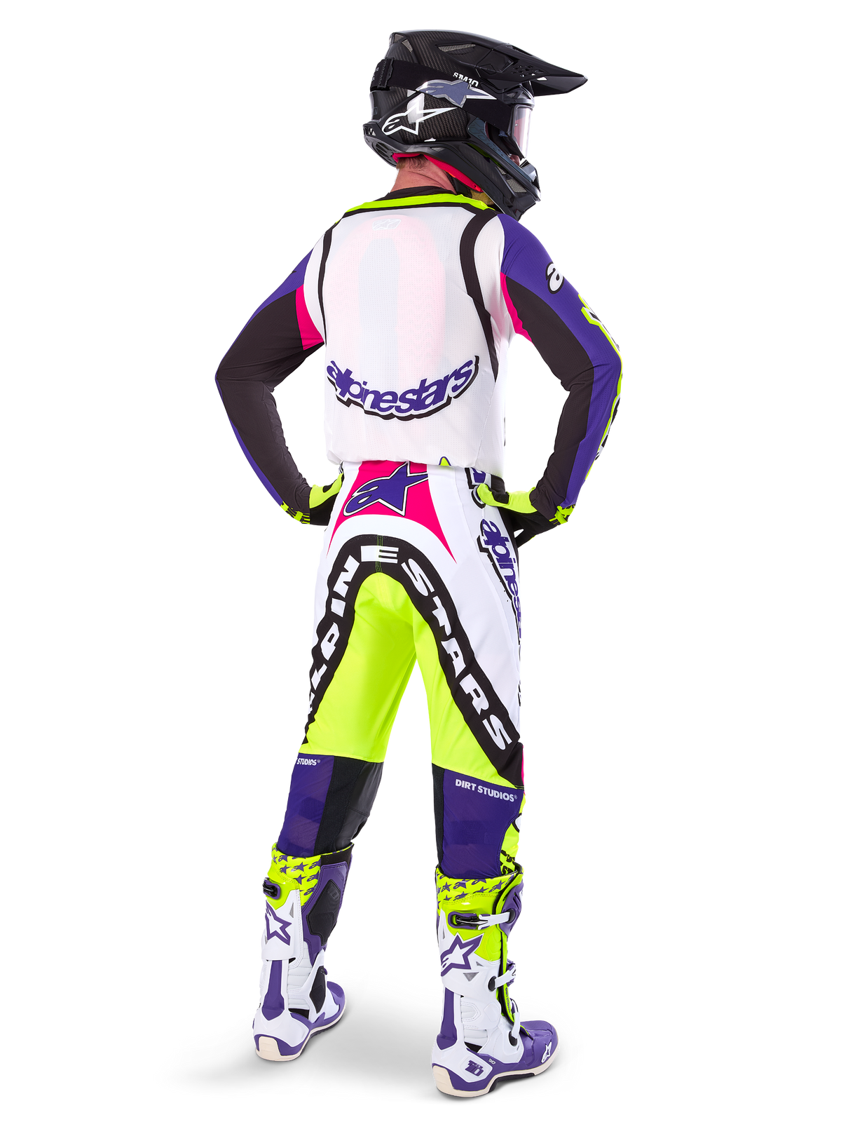 Motocyklista motocrossowy w spodniach Alpinestars Limited Edition Supertech Dirt Studios® w kolorze biało-fioletowym i żółtym fluo, widok od tyłu z dopasowaną bluzą, butami i czarnym kaskiem karbonowym, profesjonalna odzież off-road z wyraźną grafiką i neonowymi akcentami.