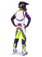 Motocyklista motocrossowy w spodniach Alpinestars Limited Edition Supertech Dirt Studios® w kolorze biało-fioletowym i żółtym fluo, widok od tyłu z dopasowaną bluzą, butami i czarnym kaskiem karbonowym, profesjonalna odzież off-road z wyraźną grafiką i neonowymi akcentami.