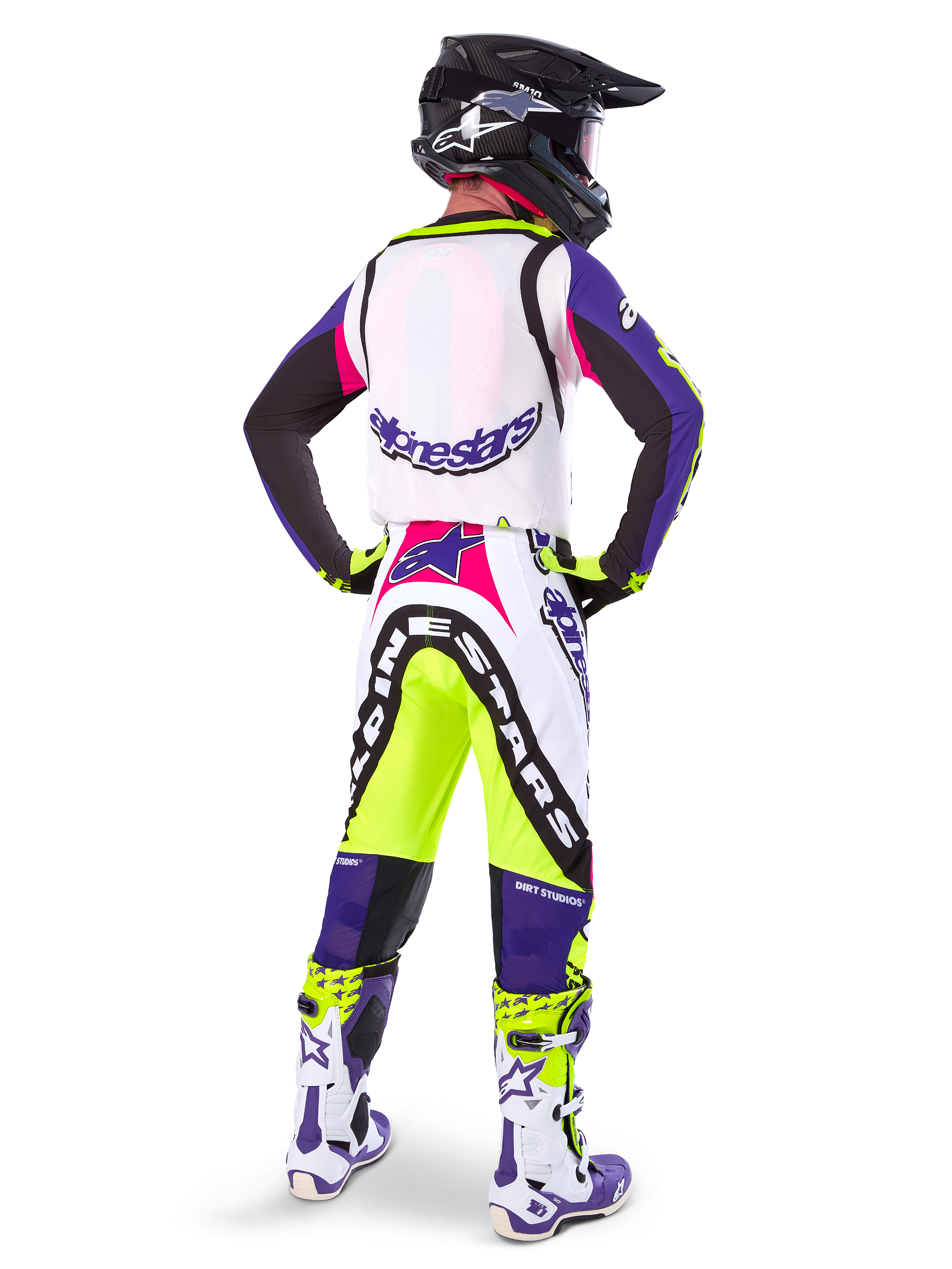 Motocyklista motocrossowy w spodniach Alpinestars Limited Edition Supertech Dirt Studios® w kolorze biało-fioletowym i żółtym fluo, widok od tyłu z dopasowaną bluzą, butami i czarnym kaskiem karbonowym, profesjonalna odzież off-road z wyraźną grafiką i neonowymi akcentami.