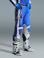 Alpinestars 2026 Racer Air Portl Pants, spodnie motocrossowe, niebiesko-białe, noszone z pasującymi niebieskimi rękawicami i biało-czarnymi butami motocrossowymi, z dużymi białymi logotypami Alpinestars, jasnobrązowymi skórzanymi panelami po wewnętrznej stronie kolan i czarną magnetyczną klamrą w pasie.