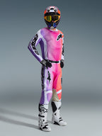 Motocyklista motocrossowy w spodniach Alpinestars 2026 Racer Air Portl Multicolor, z jaskrawym różowym, fioletowym i pomarańczowym gradientem, pasująca koszulka, biało-czarne buty motocrossowe i kask z żółtymi goglami, kompletny strój do wyścigów terenowych