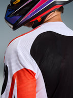 Alpinestars 2026 Racer Air Portl Jersey, koszulka motocrossowa, wielokolorowa z białymi, czarnymi i czerwonymi panelami, zbliżenie na perforowaną konstrukcję z siatki na ramieniu i plecach zapewniającą wentylację, noszona z kolorowym kaskiem motocrossowym