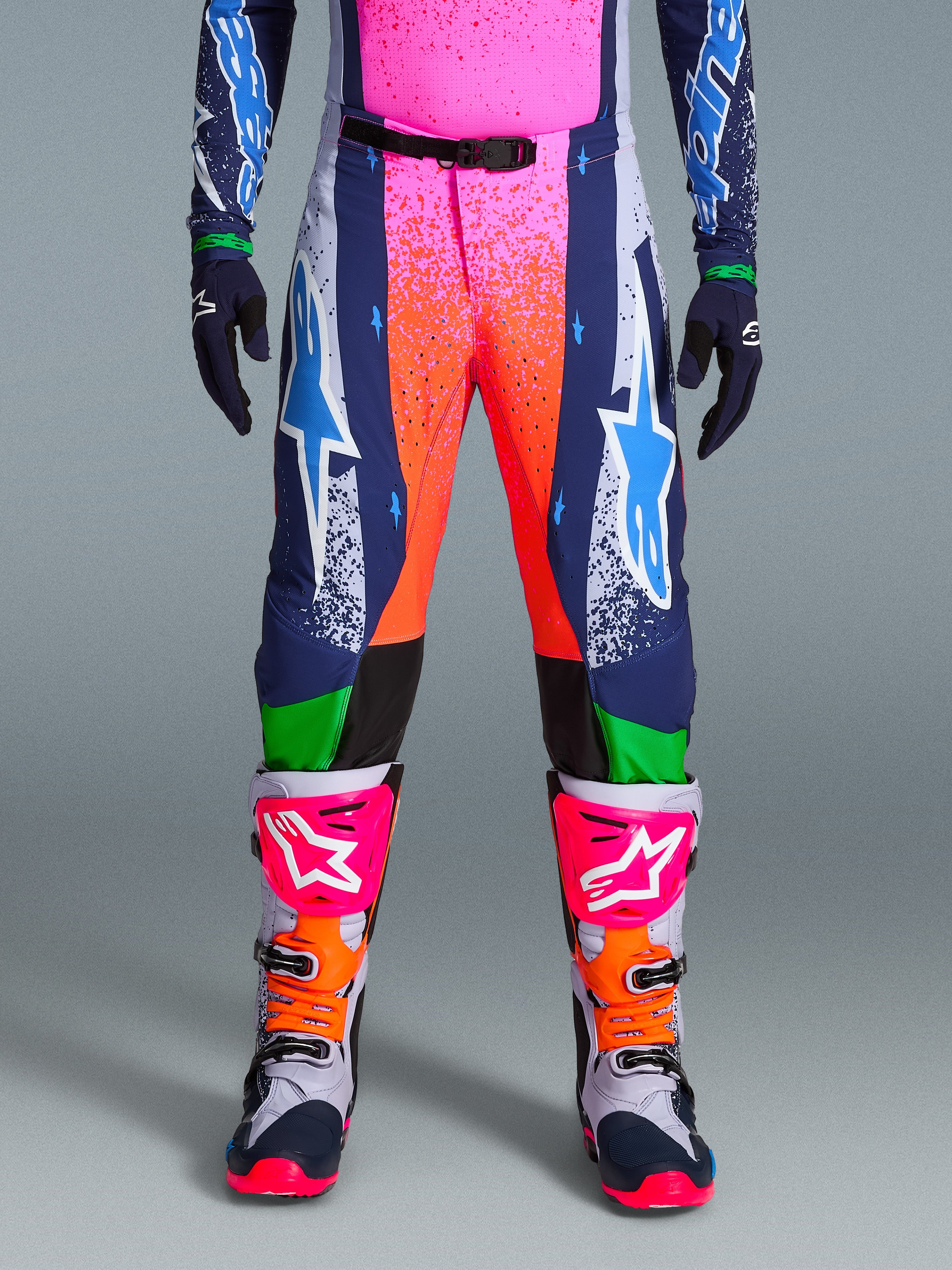 Alpinestars Limited Edition Supertech Records Pants, spodnie motocrossowe, wielokolorowe z intensywnymi różowymi i pomarańczowymi grafikami typu splash, granatowe panele z niebieskimi logo gwiazdy i zielonymi akcentami, noszone z pasującą koszulką oraz szarymi, różowymi i pomarańczowymi butami motocrossowymi