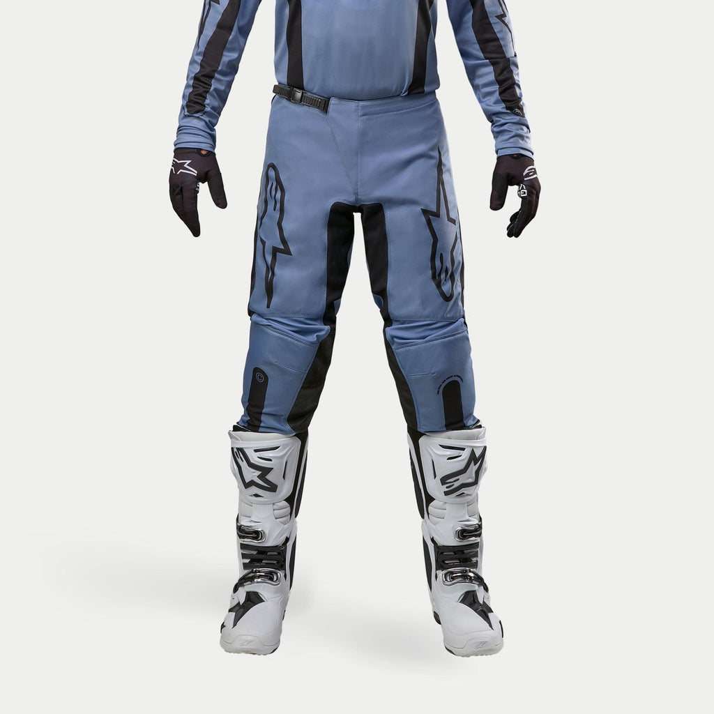 Alpinestars 2024 Fluid Lurv Pants, spodnie motocrossowe, jasnoniebieskie i czarne, noszone z biało-czarnymi butami wyścigowymi i dopasowanymi rękawicami, z wyraźnymi czarnymi logo gwiazdy na udach, ergonomiczna konstrukcja zapewniająca wydajność w terenie