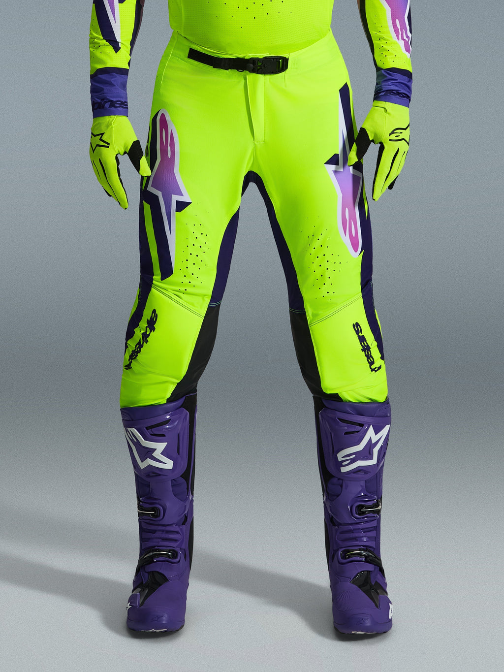 Alpinestars 2026 Supertech Vista Pants, spodnie motocrossowe, fluorescencyjny żółty, fioletowy i czarny, noszone z pasującymi rękawicami i fioletowymi butami, z dużymi gradientowymi logotypami na udach, strefami perforacji dla wentylacji i profesjonalnym sportowym designem do wyścigów off-road