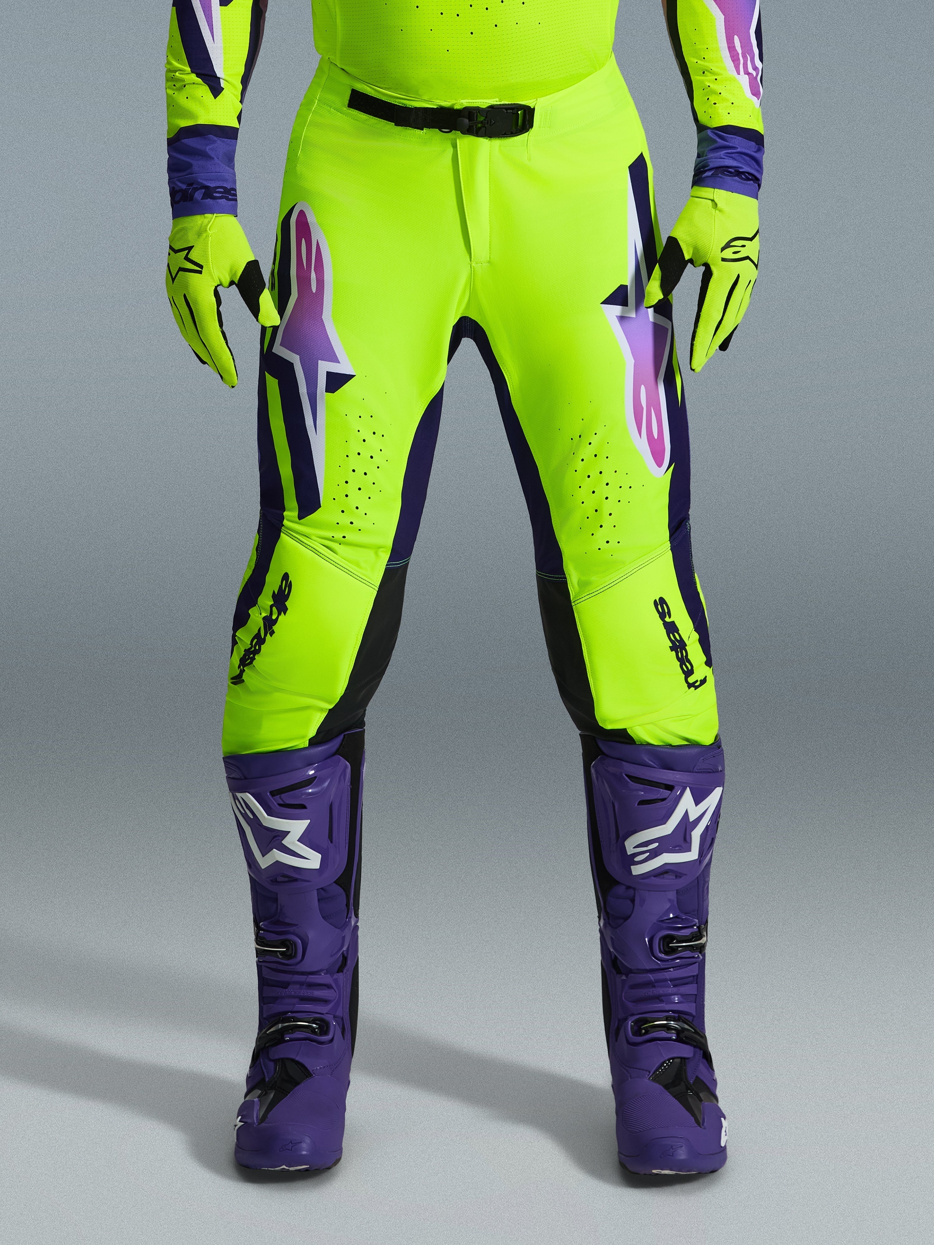 Alpinestars 2026 Supertech Vista Pants, spodnie motocrossowe, fluorescencyjny żółty, fioletowy i czarny, noszone z pasującymi rękawicami i fioletowymi butami, z dużymi gradientowymi logotypami na udach, strefami perforacji dla wentylacji i profesjonalnym sportowym designem do wyścigów off-road