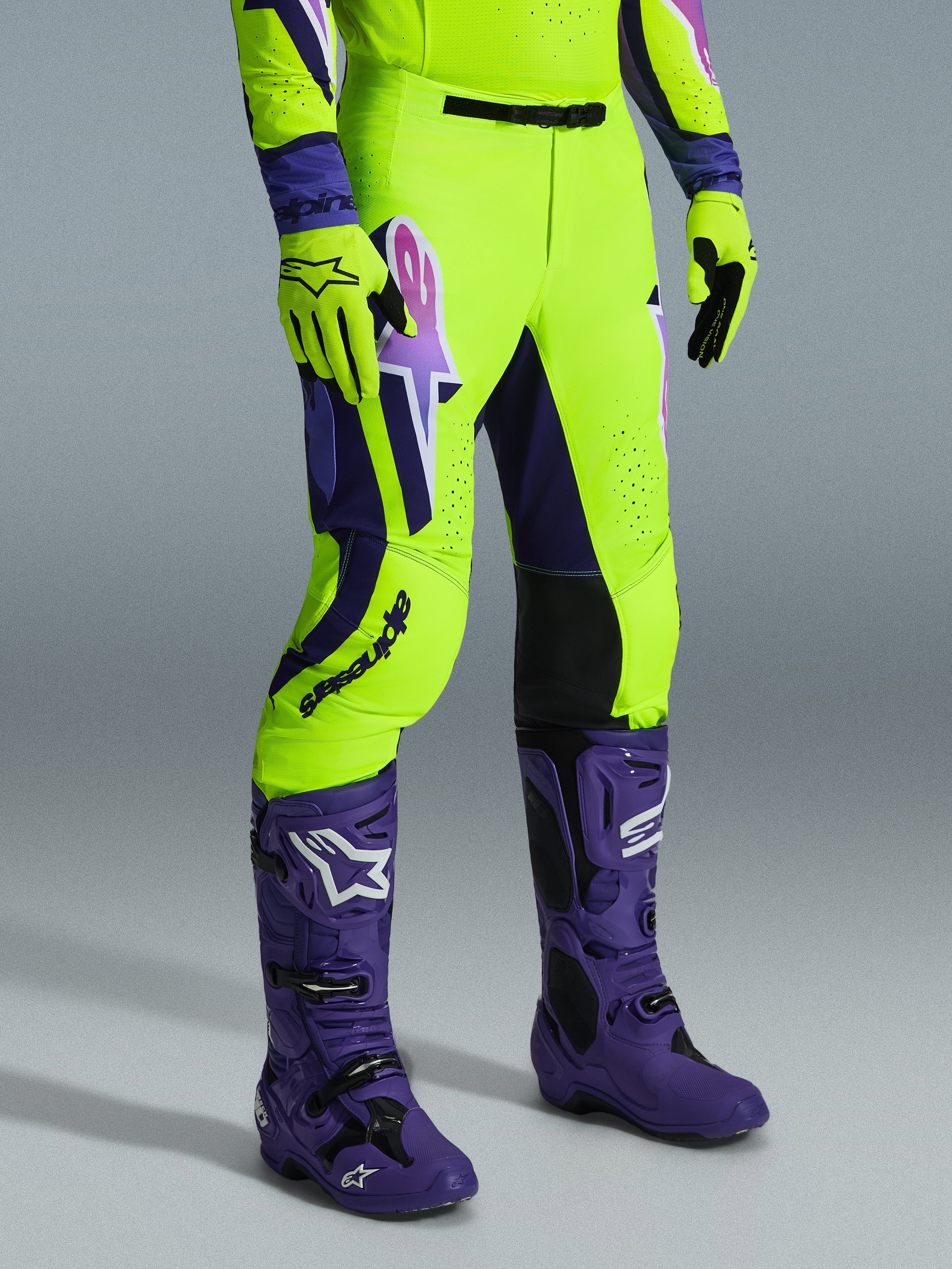 Alpinestars 2026 Supertech Vista Pants, spodnie motocrossowe, jasny neonowy żółty, fioletowy i czarny, noszone z pasującymi fioletowymi butami i żółtymi rękawicami, z wyraźnym brandingiem na udzie i sportowym, wyczynowym krojem.