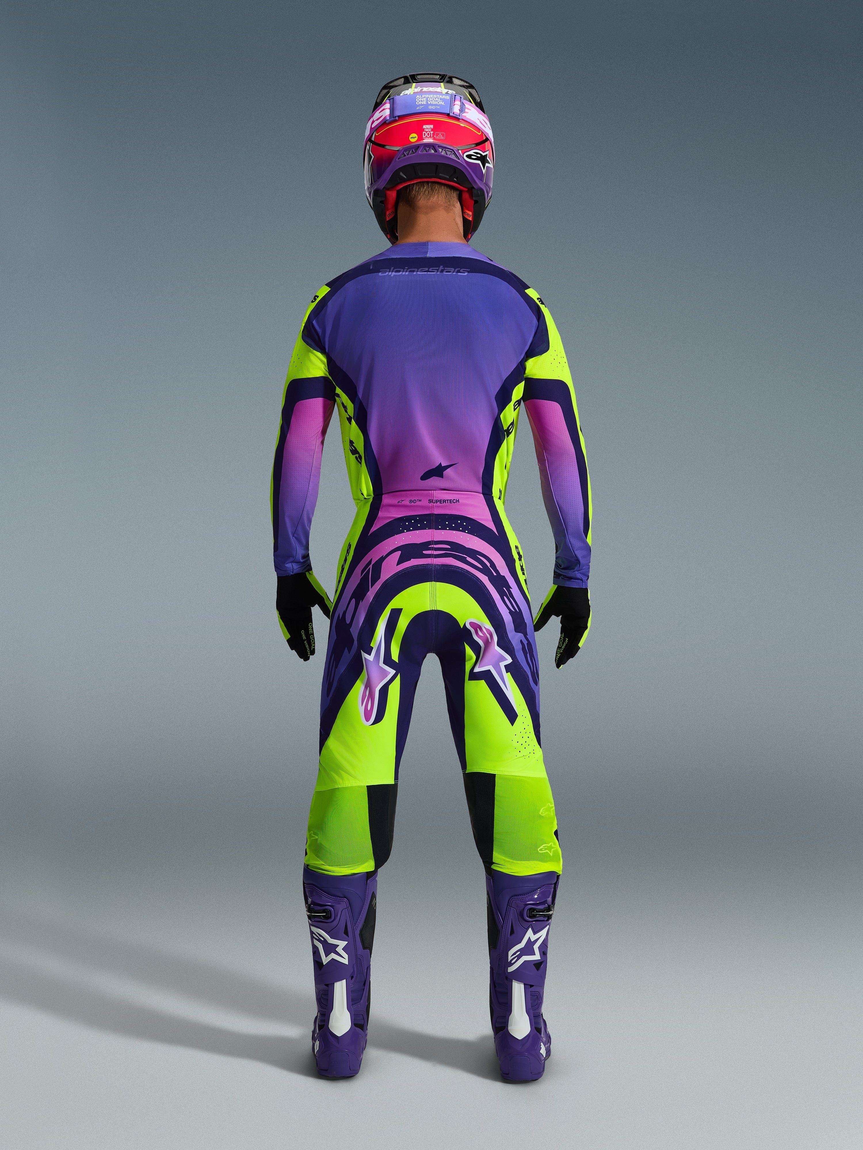 Motocyklista motocrossowy widziany od tyłu w koszulce 2026 Supertech Vista i pasujących spodniach, wyrazisty żółto-fioletowy design z różowymi gradientami, fioletowe buty z białym logo Alpinestars, czarne rękawice oraz czerwono-fioletowy kask wyścigowy, profesjonalny zestaw odzieży wyścigowej.