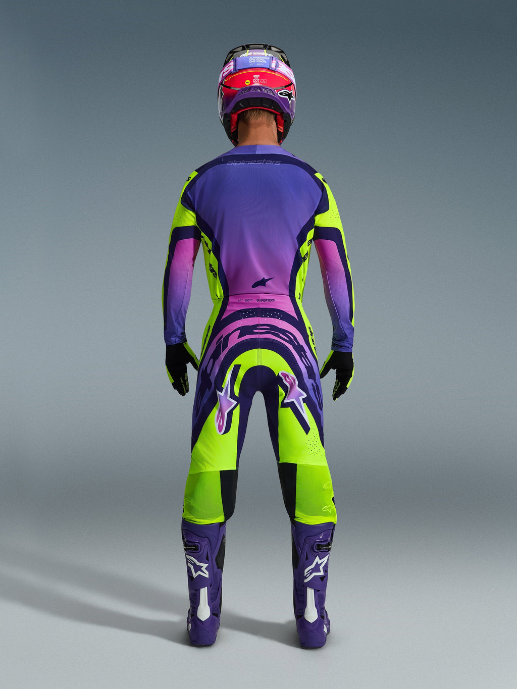 Alpinestars 2026 Supertech Vista Pants, spodnie motocrossowe, żółto-fioletowo-czarne, widok od tyłu zawodnika motocrossowego w pełnym zestawie wyścigowym z fioletową koszulką z gradientem i fioletowymi butami, wyrazisty sportowy design do profesjonalnych wyścigów off-road