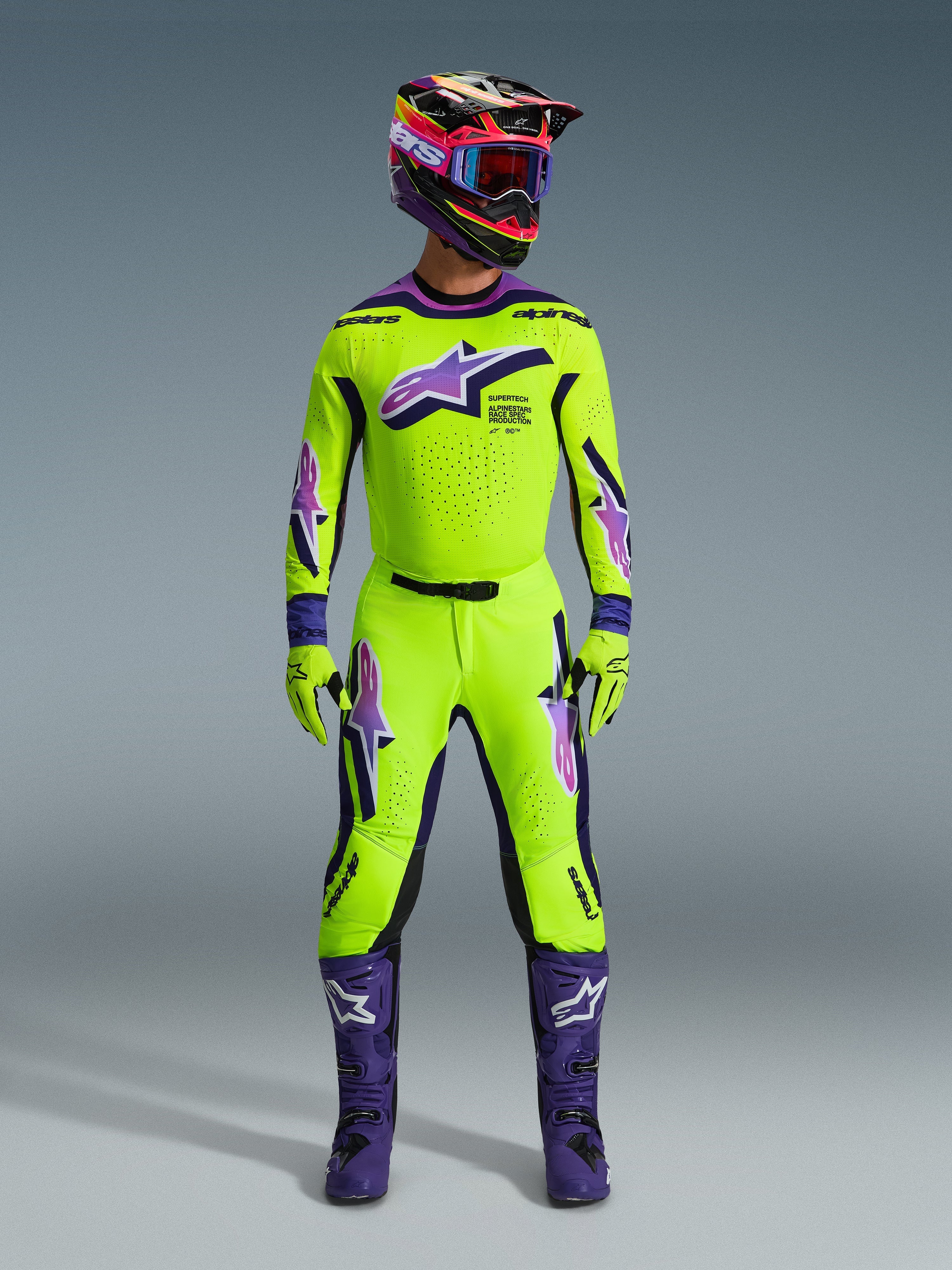 Motocyklista motocrossowy w koszulce Alpinestars 2026 Supertech Vista, jaskrawożółta z fioletowymi i czarnymi akcentami, pasujące żółto-fioletowe spodnie, fioletowe buty oraz dopasowany kask i rękawice, kompletny profesjonalny zestaw wyścigowy