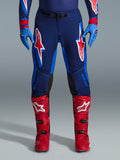 Alpinestars 2026 Supertech Vista Pants, spodnie motocrossowe, granatowe, koralowe i kobaltowe, widok z przodu na motocyklistę w profesjonalnym stroju z dopasowanymi niebieskimi rękawicami i czerwonymi butami motocrossowymi, z wyraźnym czerwono-białym logo Alpinestars na udach.