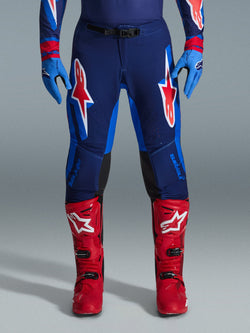 Alpinestars 2026 Supertech Vista Pants, spodnie motocrossowe, granatowe, koralowe i kobaltowe, widok z przodu na motocyklistę w profesjonalnym stroju z dopasowanymi niebieskimi rękawicami i czerwonymi butami motocrossowymi, z wyraźnym czerwono-białym logo Alpinestars na udach.