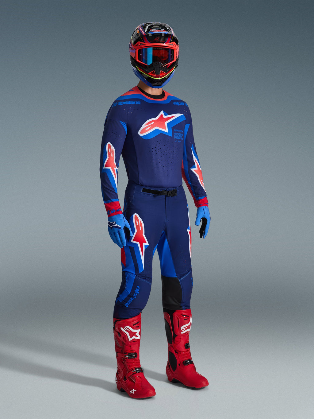 Motocyklista motocrossowy w spodniach 2026 Supertech Vista w kolorze Dark Navy Hot Coral Cobalt, dopasowana koszulka wyścigowa z długim rękawem, czerwone buty motocrossowe i kask z czerwonymi goglami, ujęcie studyjne całej sylwetki profesjonalnego stroju off-road