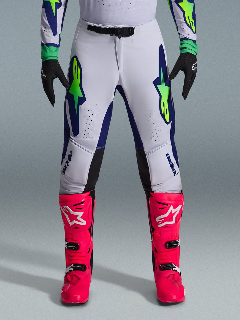 Spodnie Alpinestars Supertech Vista 2026, spodnie motocrossowe, jasnoszare z żółtymi fluorescencyjnymi i niebieskimi akcentami, zestawione z intensywnie różowymi butami i czarnymi rękawicami, posiadające wyraźne logo gwiazdy na udach oraz laserowo wycinane perforacje zapewniające przepływ powietrza.