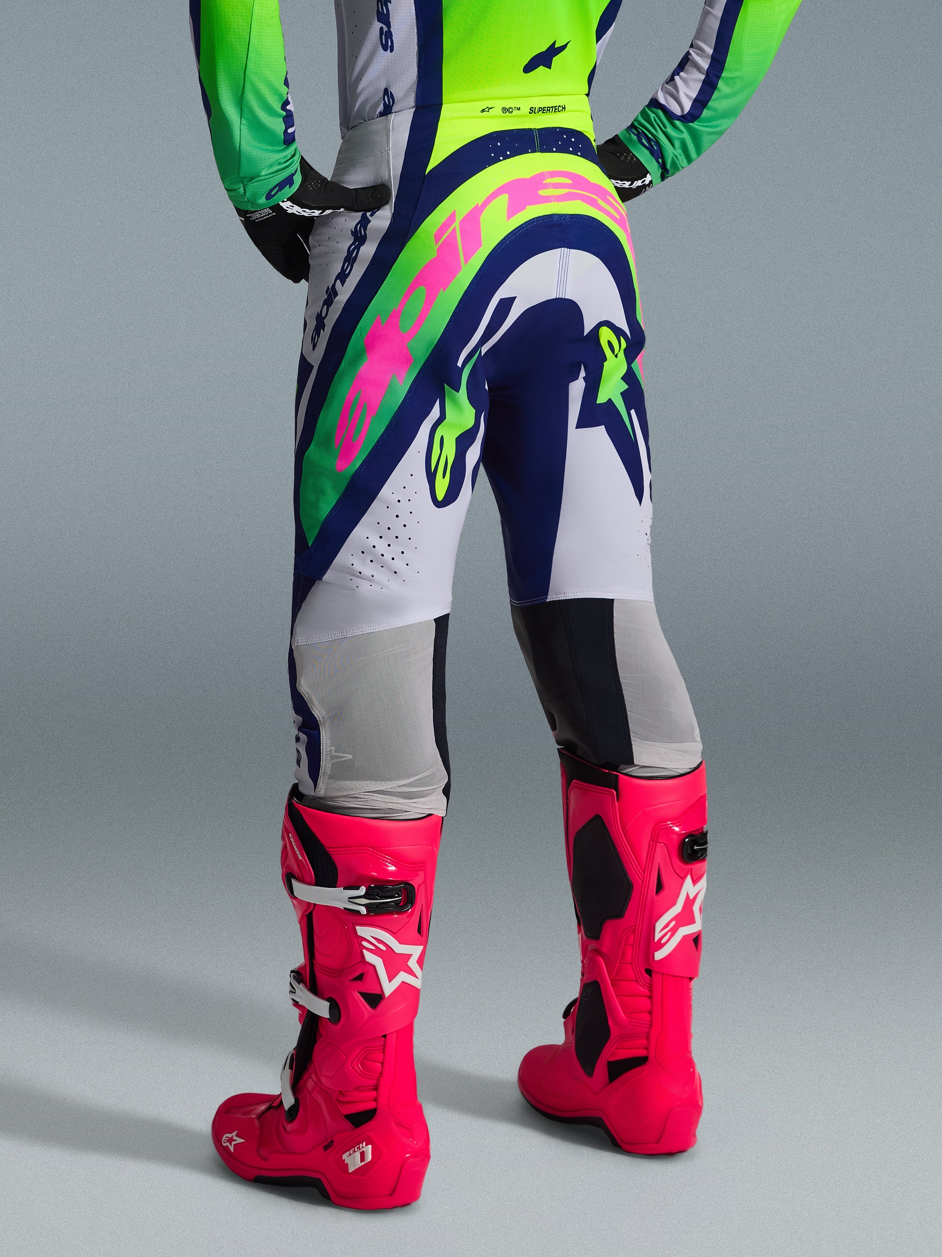 Alpinestars 2026 Supertech Vista Gear Set, widok z tyłu odzieży motocrossowej, białe spodnie z niebieskimi i limonkowymi panelami, duże różowe logo na siedzeniu, jaskraworóżowe buty Tech 10, profesjonalny sprzęt do wyścigów terenowych