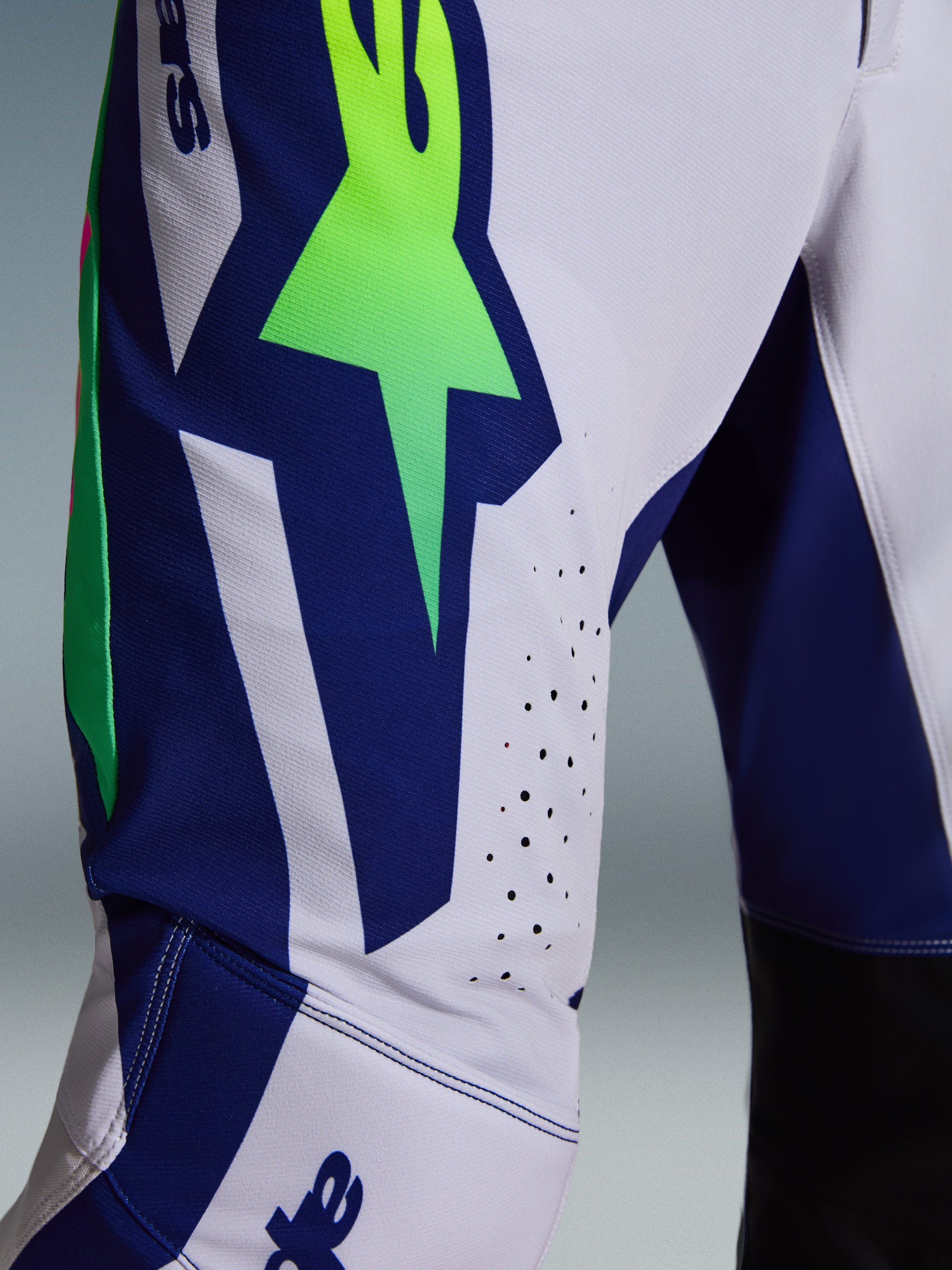 Alpinestars 2026 Supertech Vista Pants, spodnie motocrossowe, jasnoszare z żółtymi fluorescencyjnymi i niebieskimi akcentami, zbliżenie na udo z dużym logo A-star, laserowo wycinane otwory wentylacyjne, zaawansowany elastyczny materiał rip-stop