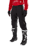 Alpinestars Techdura Pants, czarne spodnie motocyklowe off-road, zestawione z czerwoną bluzą z długim rękawem i czarno-białymi butami motocrossowymi, wytrzymała konstrukcja z elastycznego poliamidu, skórzane wzmocnienia wewnętrznej strony kolan, regulowany pasek z klamrą do jazdy enduro