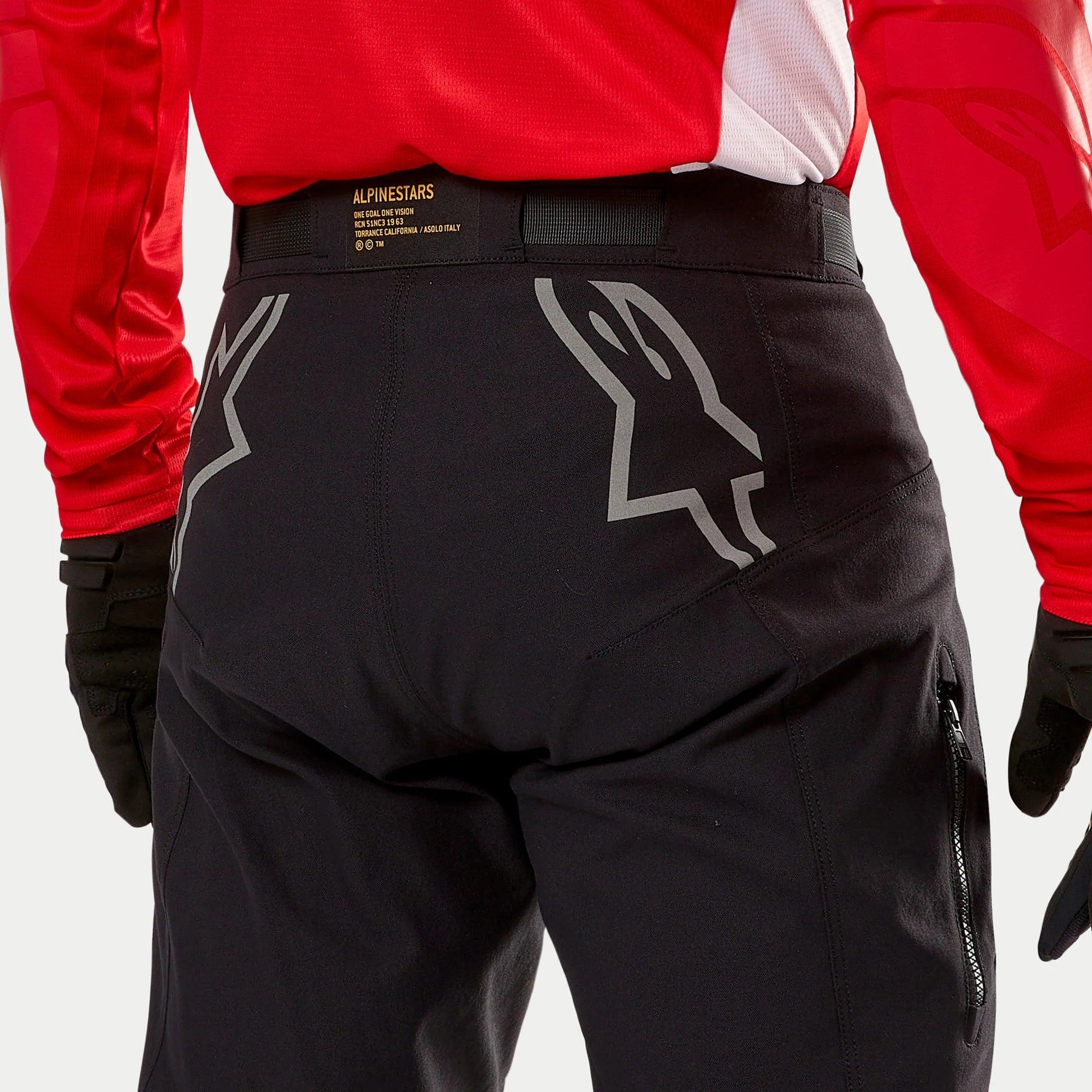 Alpinestars Techdura Pants, czarne spodnie enduro, widok z tyłu z dużymi szarymi logotypami Astars na siedzeniu, noszone z czerwoną bluzą i czarnymi rękawicami, wytrzymały materiał z bocznym zamkiem i markowym pasem