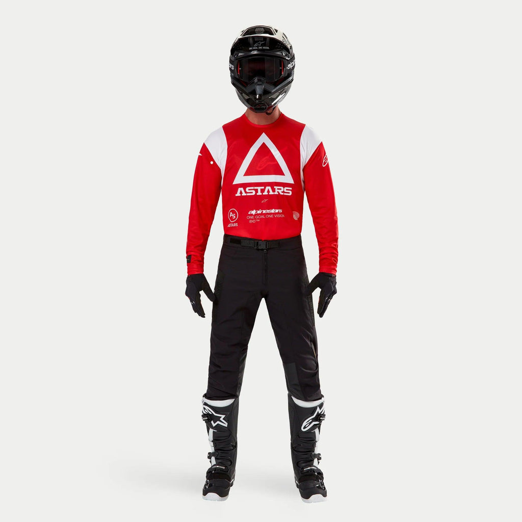 Spodnie Alpinestars Techdura, spodnie enduro i off-road, czarne, noszone z czerwono-białą bluzą Astars z długim rękawem, czarnym kaskiem i czarno-białymi butami, cechujące się wytrzymałą konstrukcją z rozciągliwego poliamidu i technicznymi panelami do jazdy terenowej.