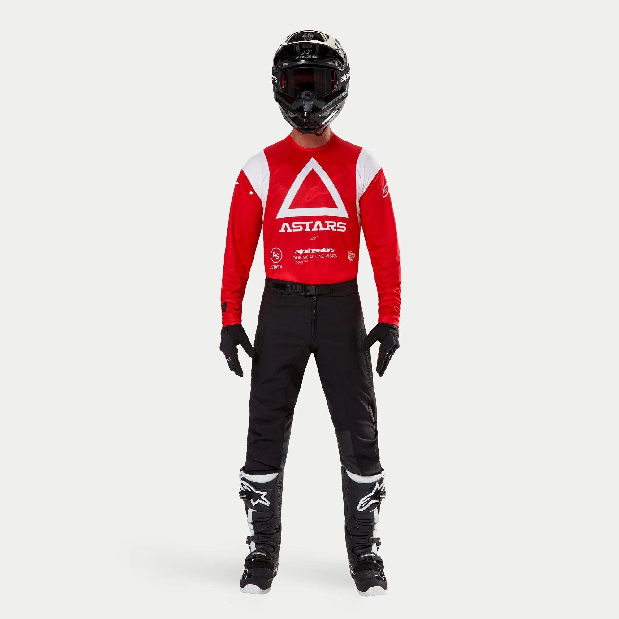 Spodnie Alpinestars Techdura, spodnie enduro i off-road, czarne, noszone z czerwono-białą bluzą Astars z długim rękawem, czarnym kaskiem i czarno-białymi butami, cechujące się wytrzymałą konstrukcją z rozciągliwego poliamidu i technicznymi panelami do jazdy terenowej.
