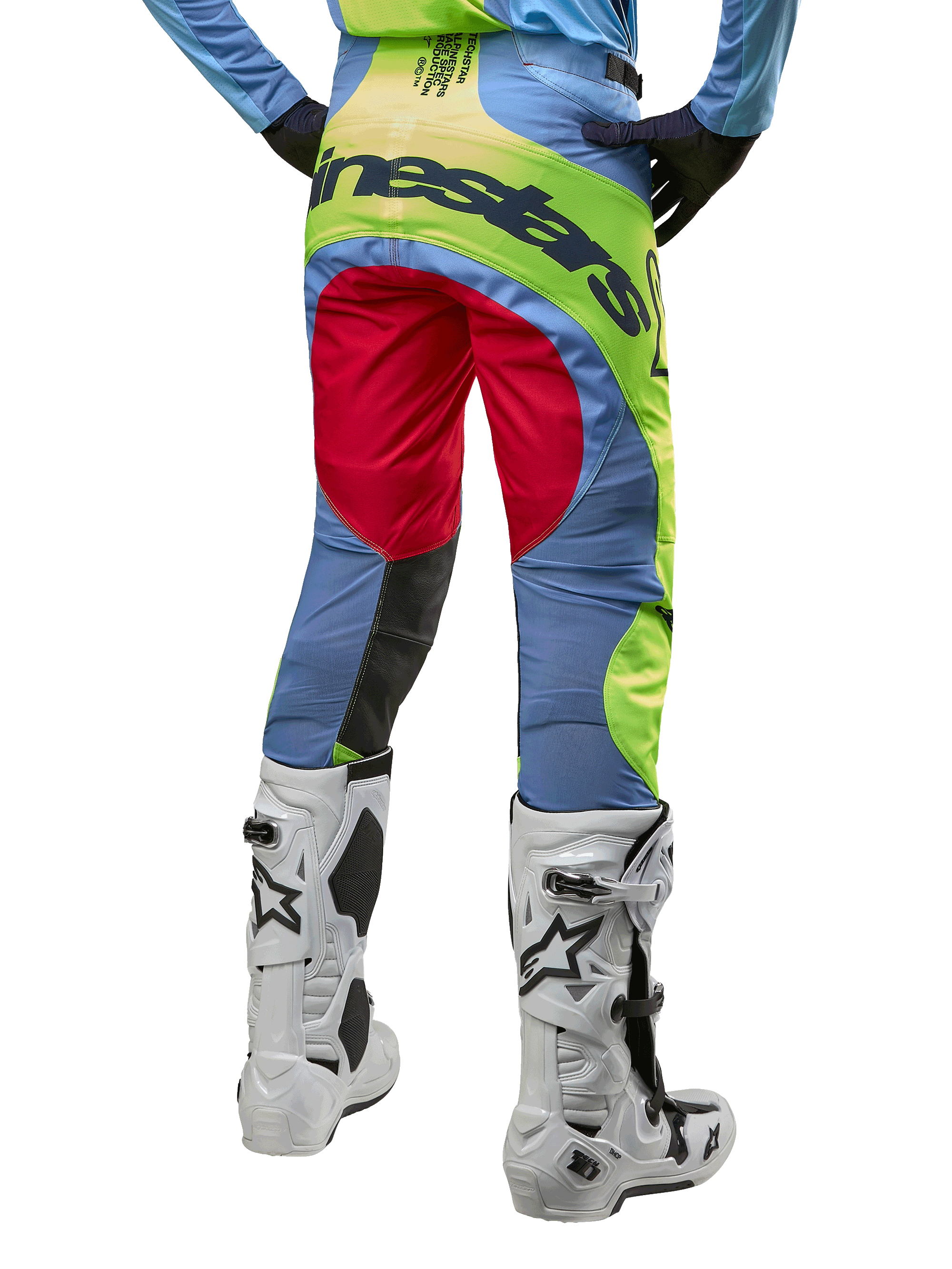 Alpinestars Techstar Ocuri Pants 2024, spodnie motocrossowe, jasnoniebieskie, neonowe żółte i czerwone, widok z tyłu w biało-czarnych butach motocrossowych, wyrazisty design color-blocking i duże logo Alpinestars na siedzeniu