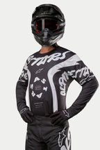 Alpinestars 2024 Racer Hana Jersey, koszulka motocrossowa, czarno-biała, z wyraźną białą grafiką tekstową i logo Alpinestars, noszona przez zawodnika w czarnym kasku i goglach, profesjonalna odzież do wyścigów terenowych