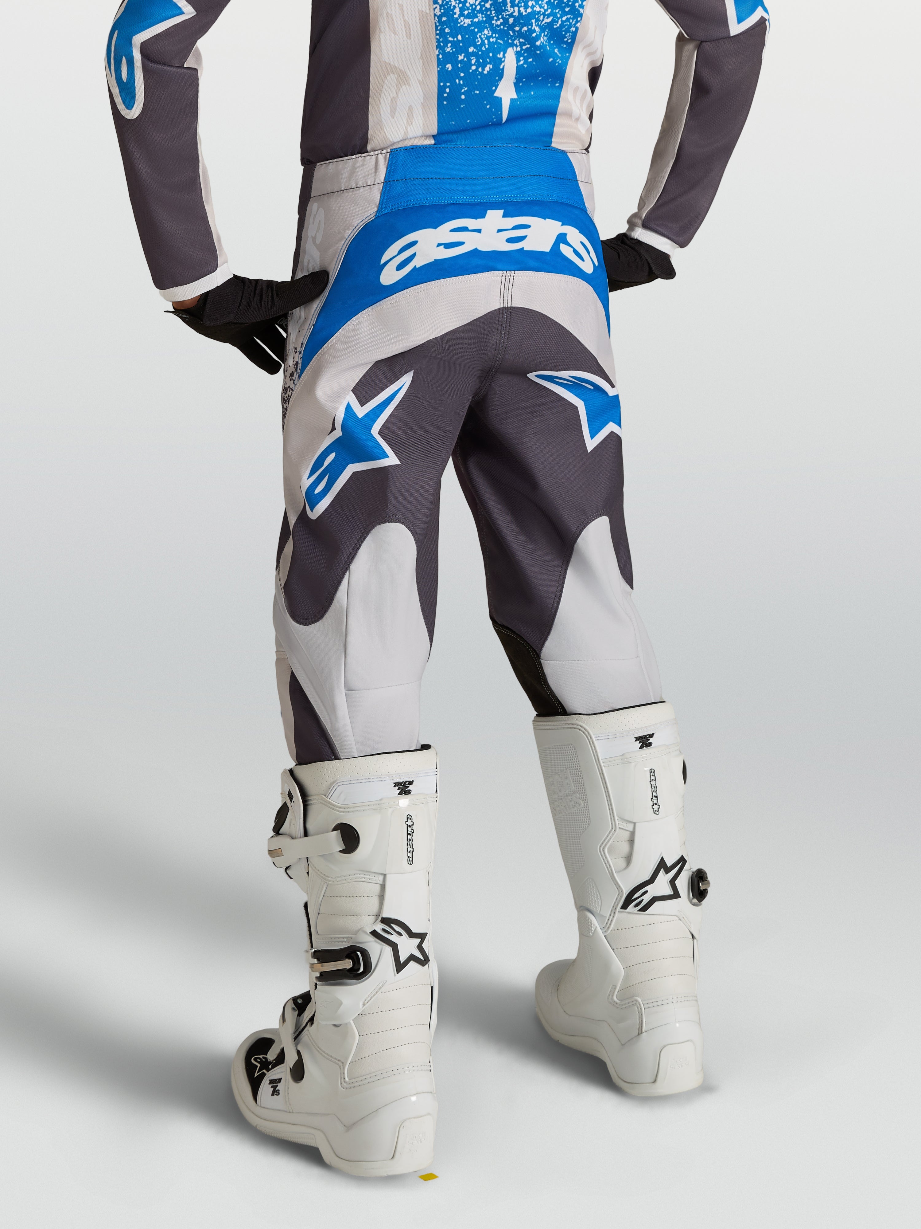Alpinestars Spodnie Fluid Drag Młodzieżowe, szare i niebieskie spodnie motocrossowe, widok od tyłu na ridera w białych butach.