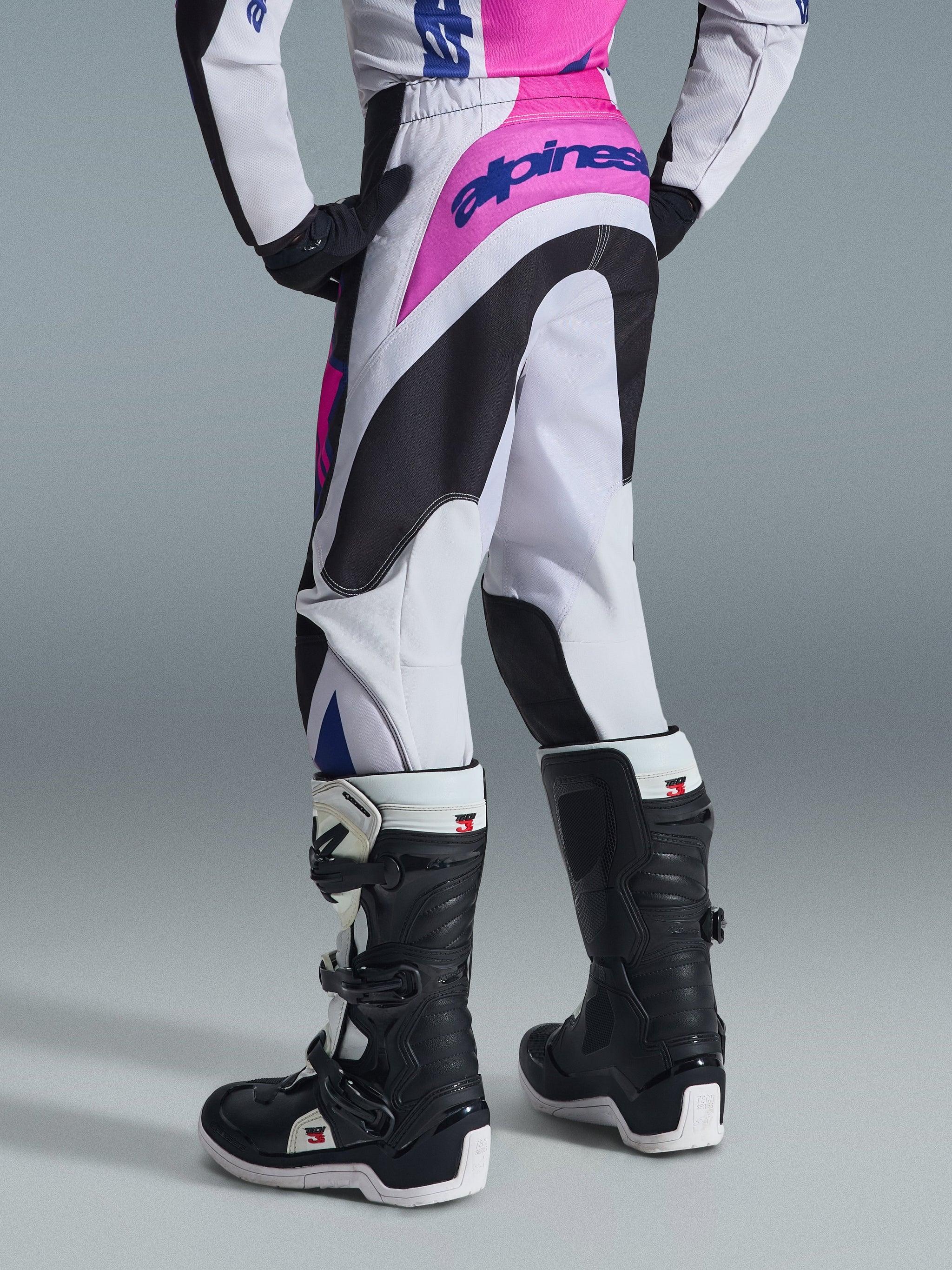 Alpinestars 2026 Youth Fluid Grid Pants, Motocross Pants, Black Light Gray and Purple, rear view showing bold pink branding and ergonomic black panels, worn with black and white Tech 3 boots, high-performance youth off-road gear