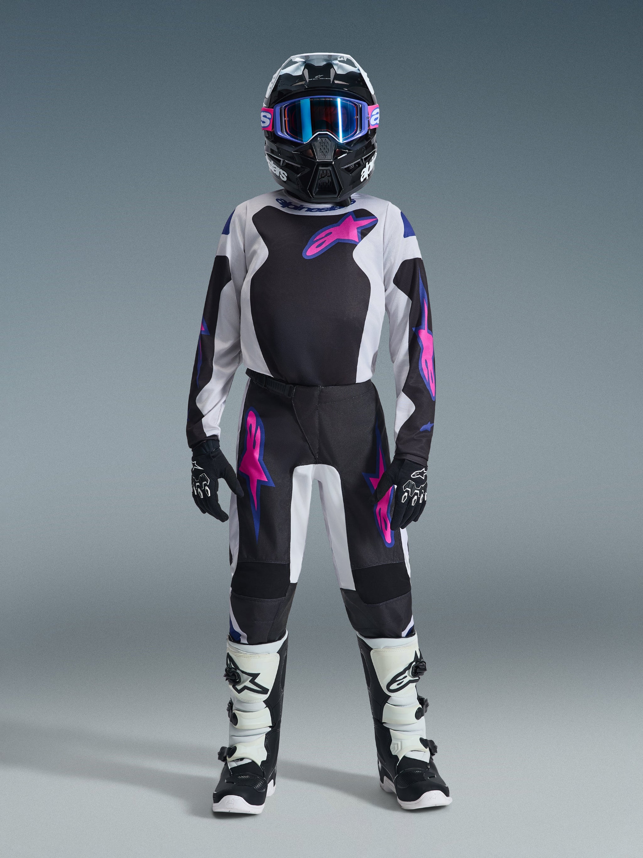 Youth motocross rider wearing Alpinestars 2026 Youth Fluid Grid Pants in Black, Light Gray, and Purple, matching long-sleeve jersey, black helmet with pink and purple goggles, white and black boots, and black gloves, full coordinated gear set for off-road riding.