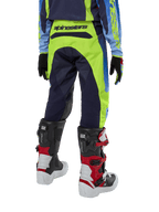 Alpinestars Youth 2024 Racer Hoen Pants, spodnie motocrossowe, żółte fluo i granatowe, widok z tyłu prezentujący wyraźne logo Alpinestars na pasie, noszone z czarno-czerwonymi butami Tech Series i pasującą koszulką, wytrzymały sportowy krój do jazdy terenowej dla młodzieży