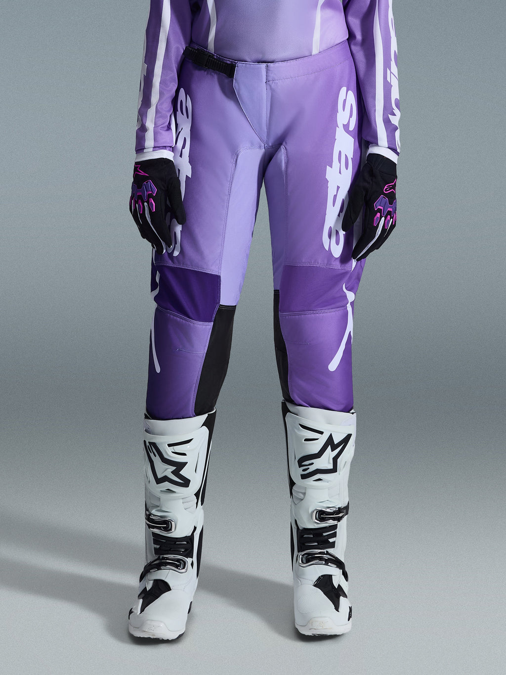 Alpinestars Stella Fluid Apex Pants 2026, damskie spodnie motocrossowe, fioletowe i białe, noszone z czarno-fioletowymi rękawicami i biało-czarnymi butami, z wyraźnym białym logo na nogawce, ergonomiczny sportowy krój do wyścigów terenowych