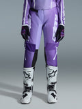 Alpinestars Stella Fluid Apex Pants 2026, damskie spodnie motocrossowe, fioletowe i białe, noszone z czarno-fioletowymi rękawicami i biało-czarnymi butami, z wyraźnym białym logo na nogawce, ergonomiczny sportowy krój do wyścigów terenowych