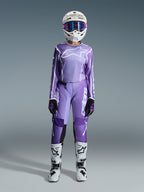 Zawodniczka motocrossowa w zestawie Alpinestars 2026 Stella Fluid Apex, fioletowo-biała koszulka i pasujące spodnie, biały kask z różowymi goglami oraz biało-czarne buty off-road, kompletny zestaw wyścigowy do damskiego motocrossu.
