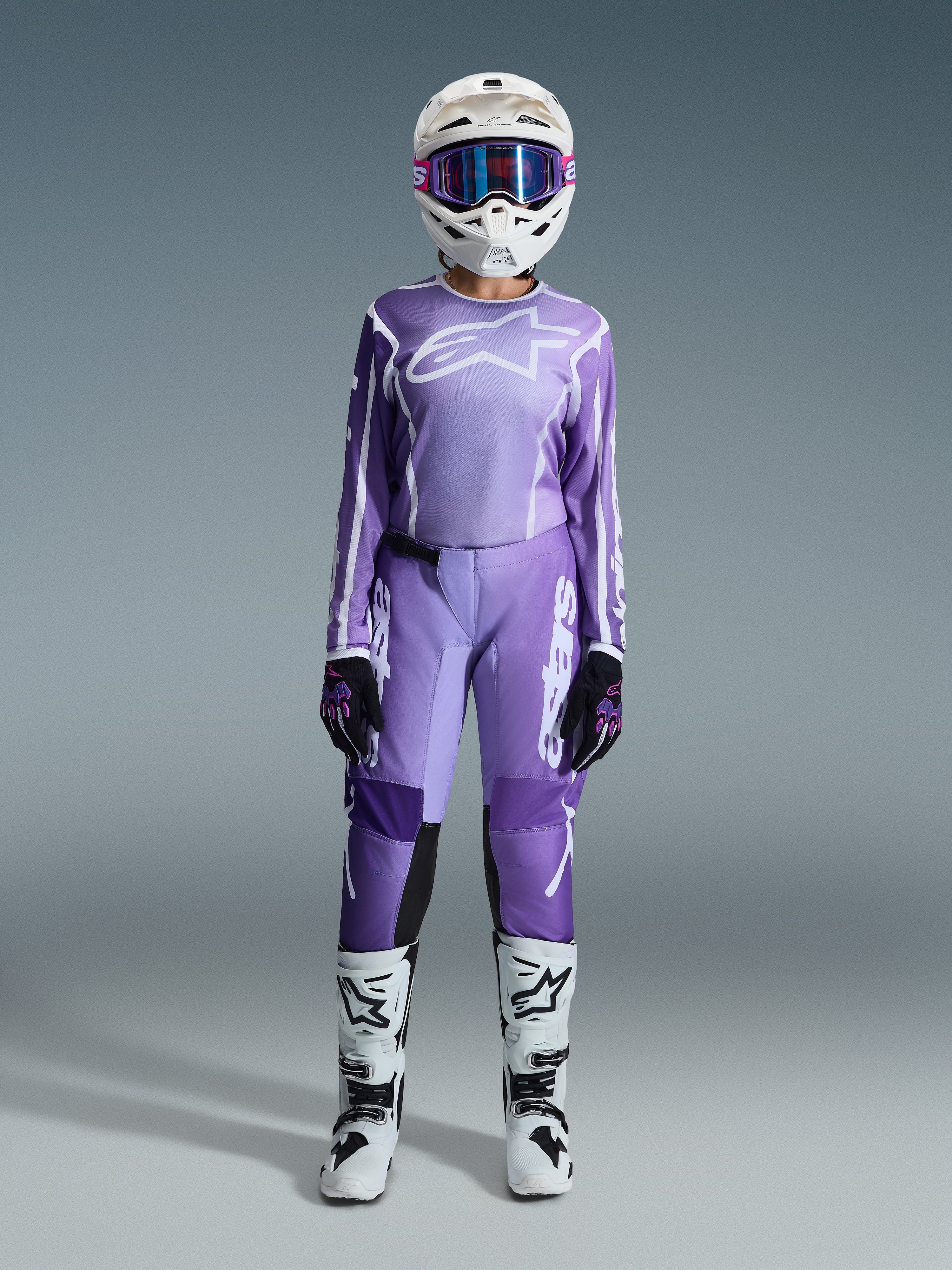 Zawodniczka motocrossowa w zestawie Alpinestars 2026 Stella Fluid Apex, fioletowo-biała koszulka i pasujące spodnie, biały kask z różowymi goglami oraz biało-czarne buty off-road, kompletny zestaw wyścigowy do damskiego motocrossu.